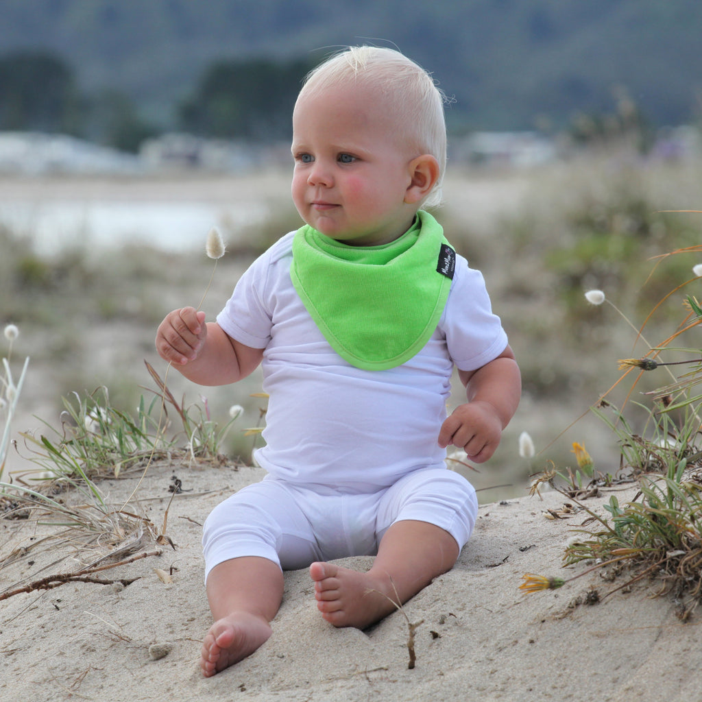 Baby sitting on sand wearing a white outfit and green bib 