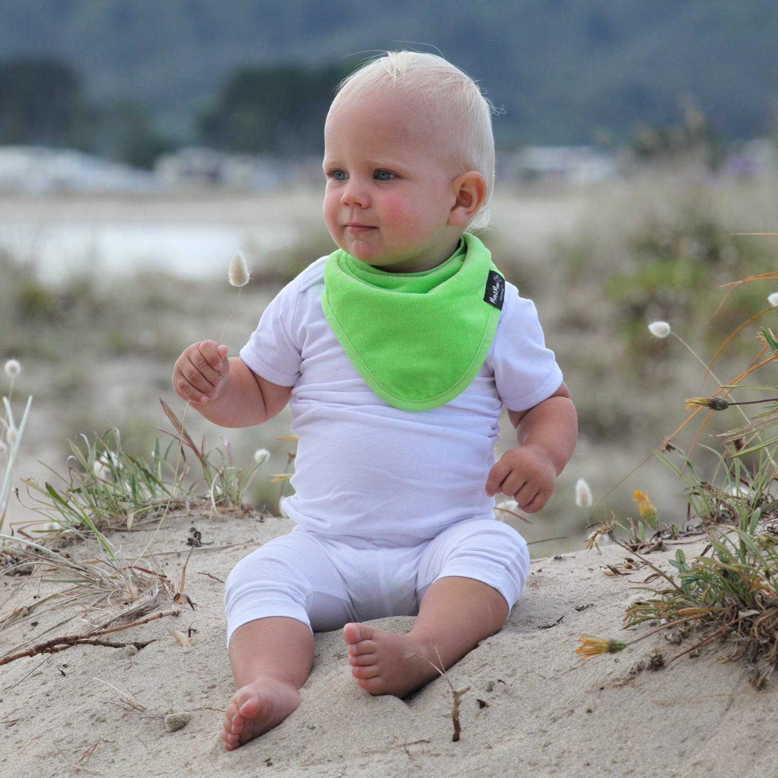 Baby sitting on sand wearing a white outfit and green bib 