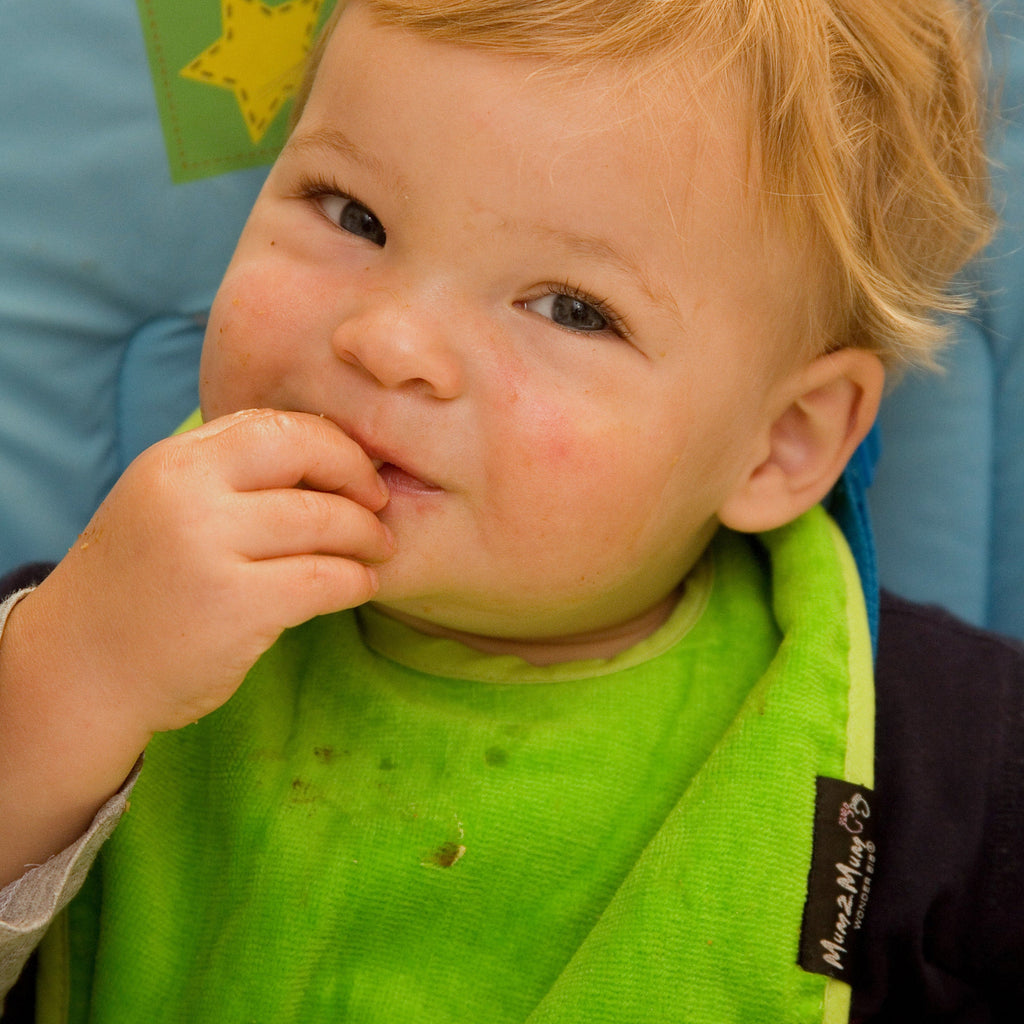 A close-up of a young child wearing a bright green bib with a small black brand tag that reads 'Mum 2 Mum'. The child is sitting in a blue high chair and has a hand near their mouth.