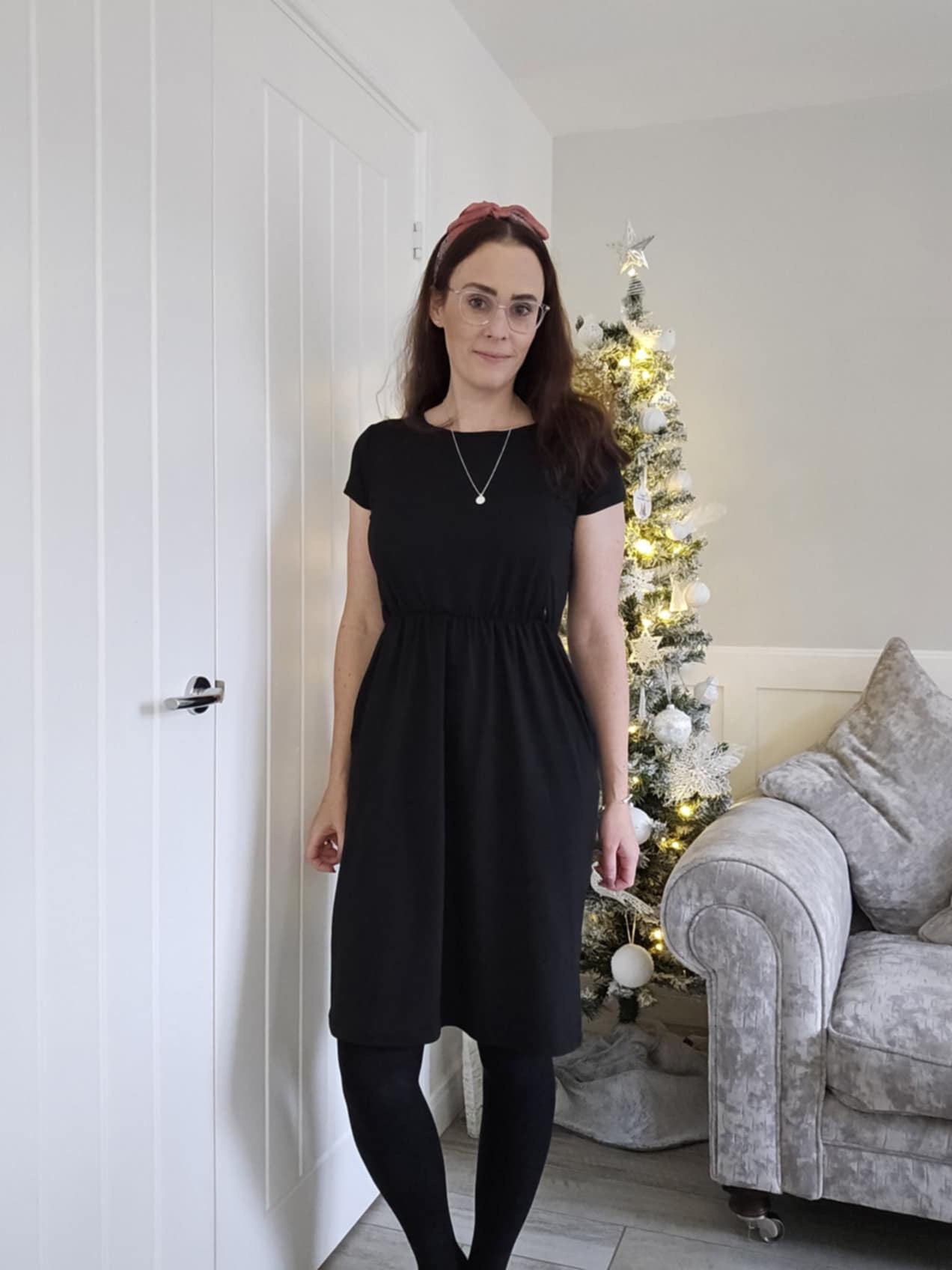 Woman in a black dress standing in a room with a Christmas tree and gray sofa.