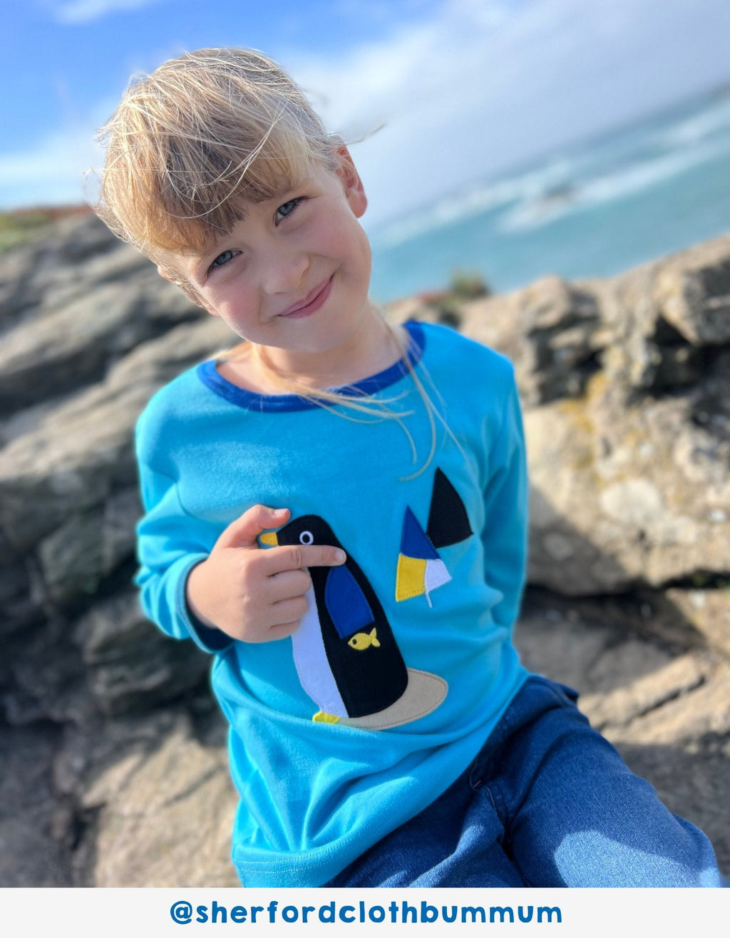 Child wearing a blue shirt with a penguin design, sitting on rocks by the sea.
