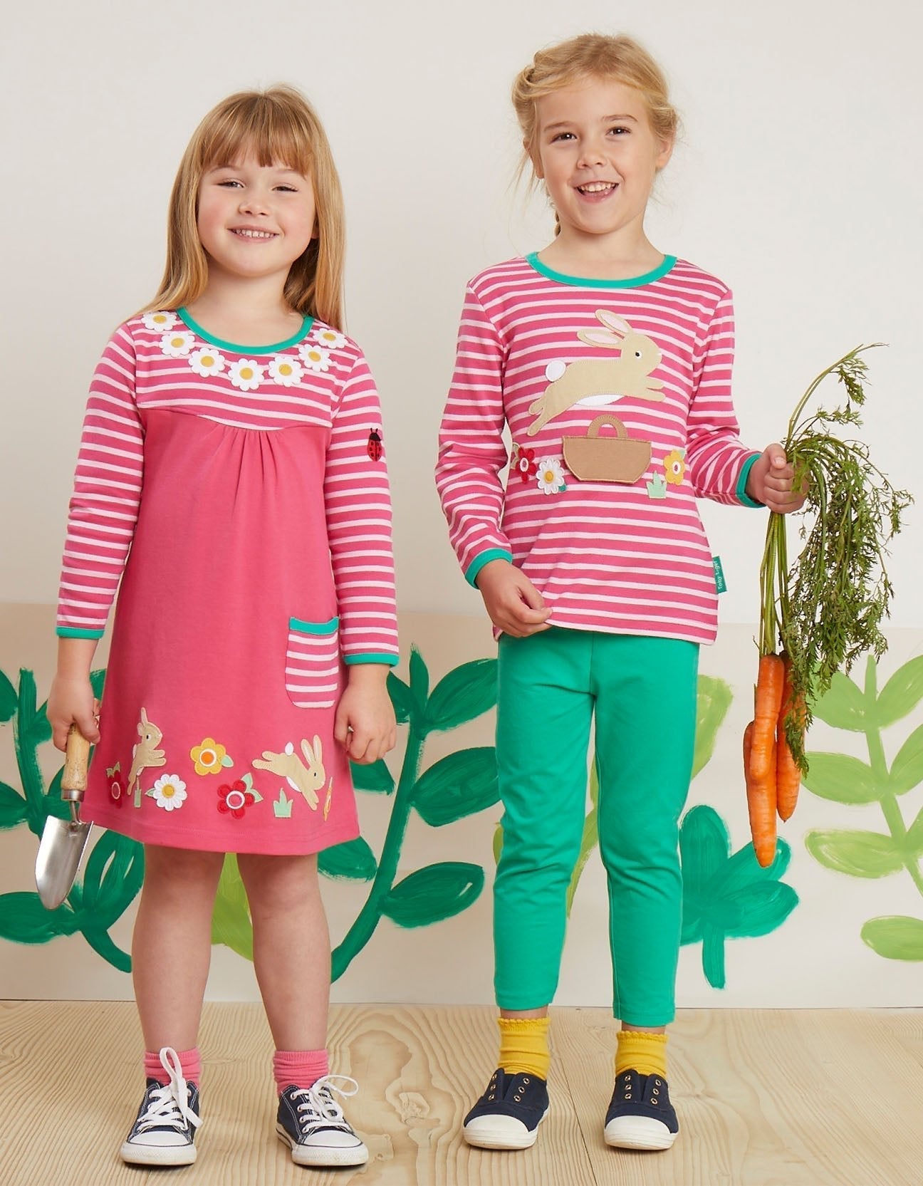 Two children wearing pink and green outfits with animal designs against a leafy background.