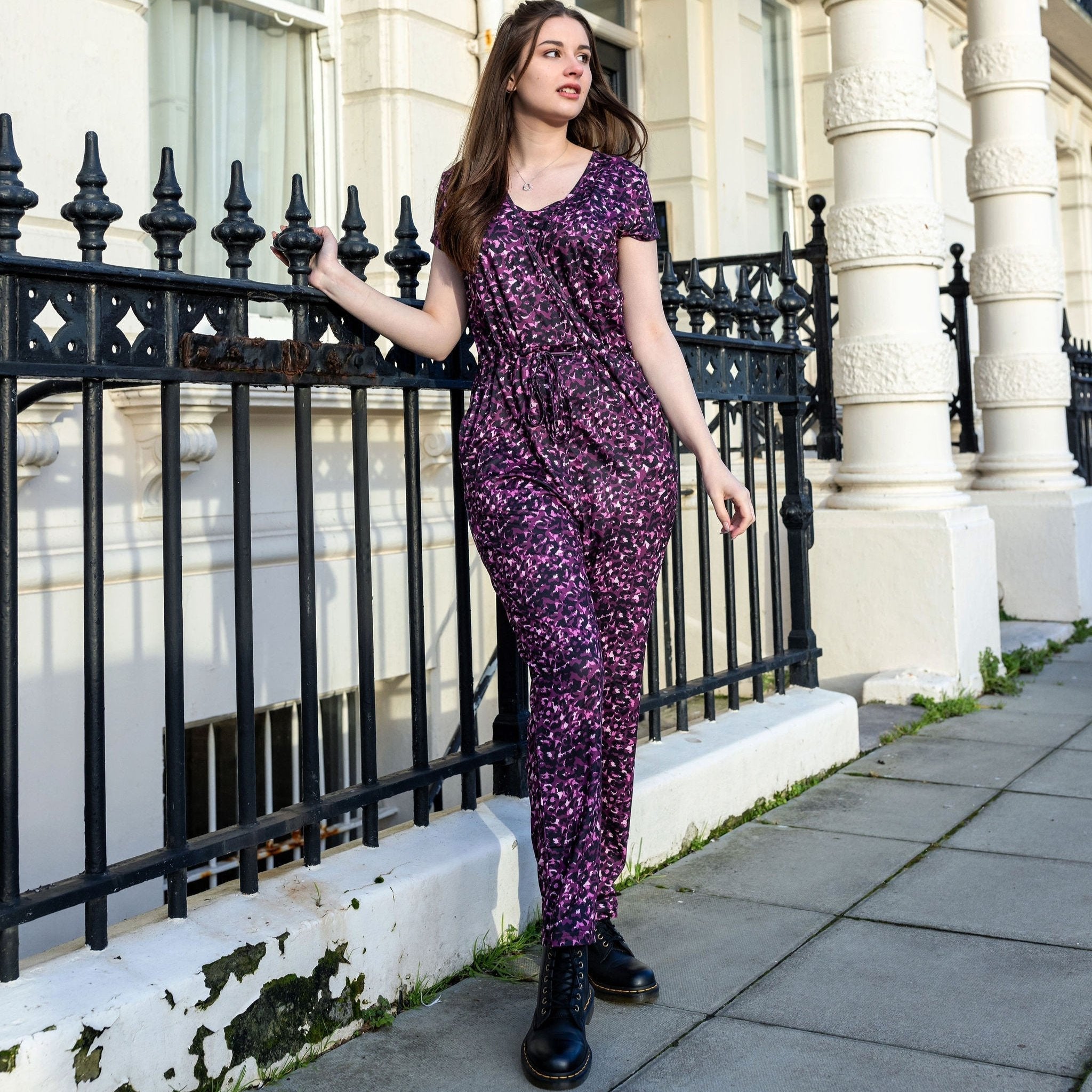 Woman in a purple floral jumpsuit standing on a sidewalk next to a black iron fence.