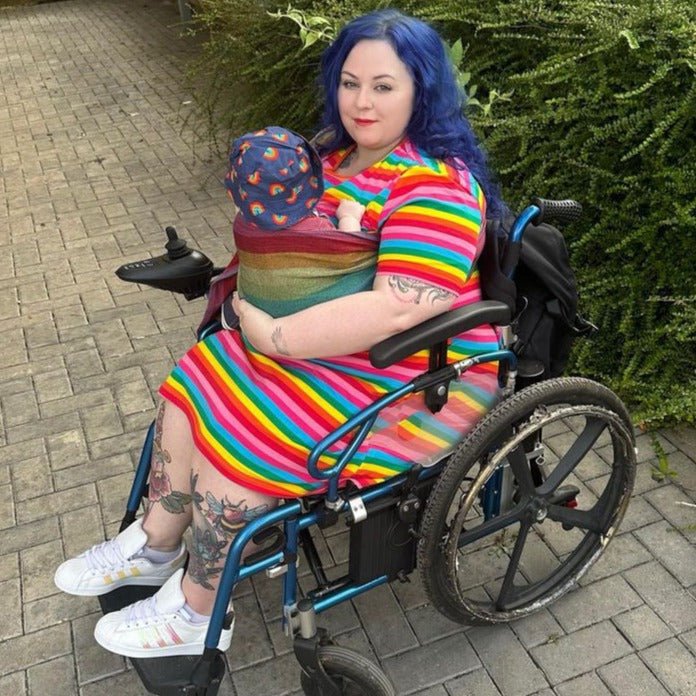 Person in a wheelchair wearing a rainbow dress, with a baby outdoors.